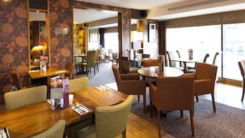 Espaço de refeições de hotel com várias mesas e cadeiras, um espelho grande e vista para a rua.