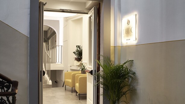 Un pasillo con una planta en maceta, un mostrador de recepción y una escalera que lleva a una habitación con cama y escritorio.