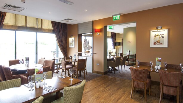 A hotel dining area with tables, chairs, and a view of the outdoors. - Premier Inn London Stansted Airport (Bishop's Stortford)