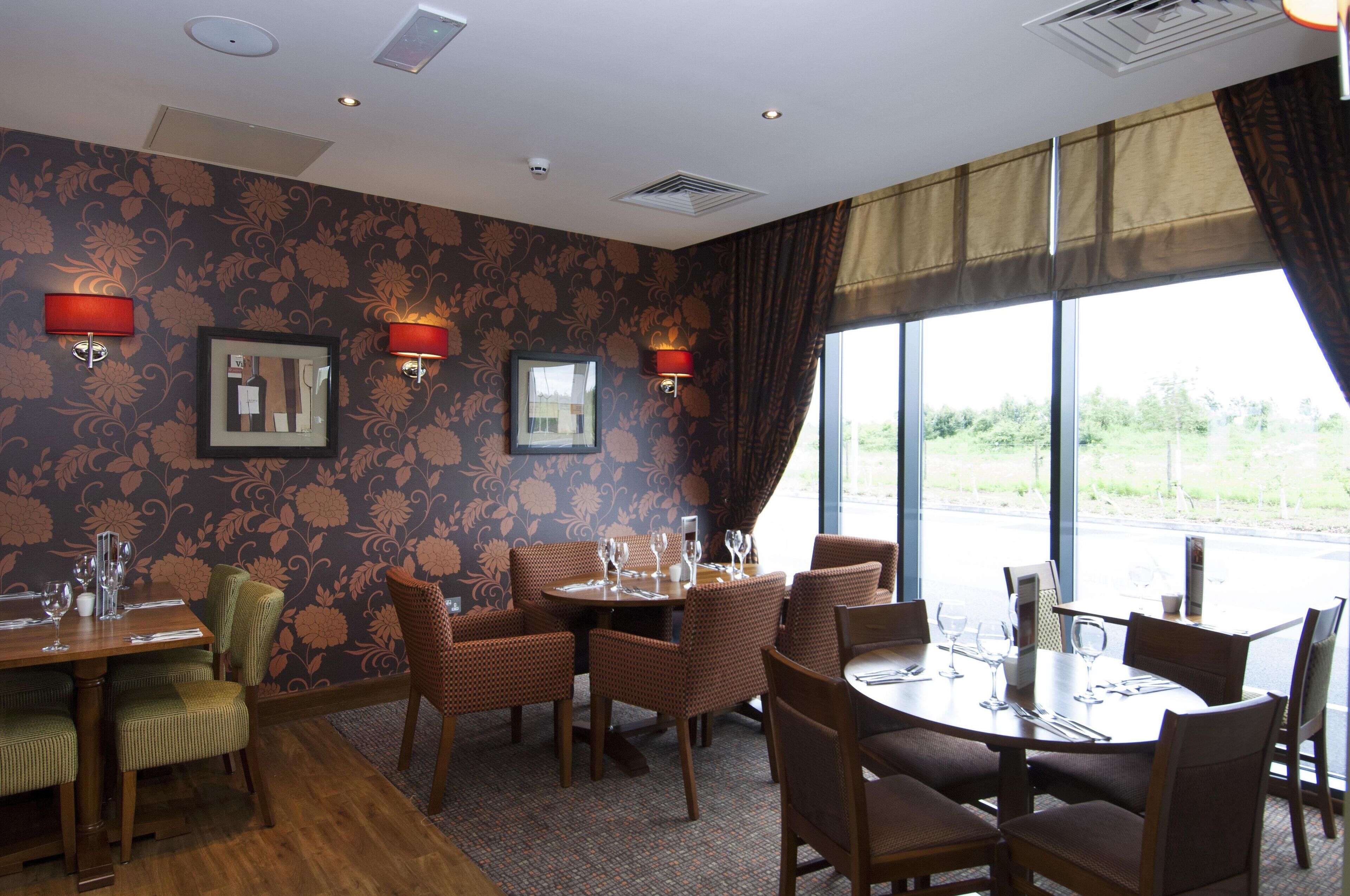 A dining area with patterned wallpaper, wall-mounted lamps, and large windows with curtains.