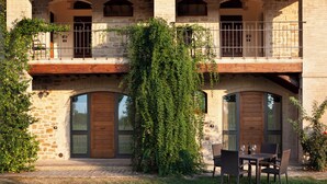 A stone building with a wooden balcony, a dining area with chairs and a table, and a large tree in front. - Valle di Assisi Country Apartments (Assisi)