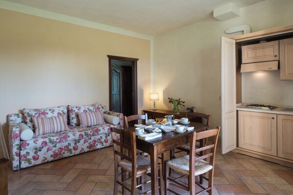 Una sala de estar con un sofá floral, una mesa de comedor de madera y sillas, una cocina con armarios de madera y un suelo de baldosas.