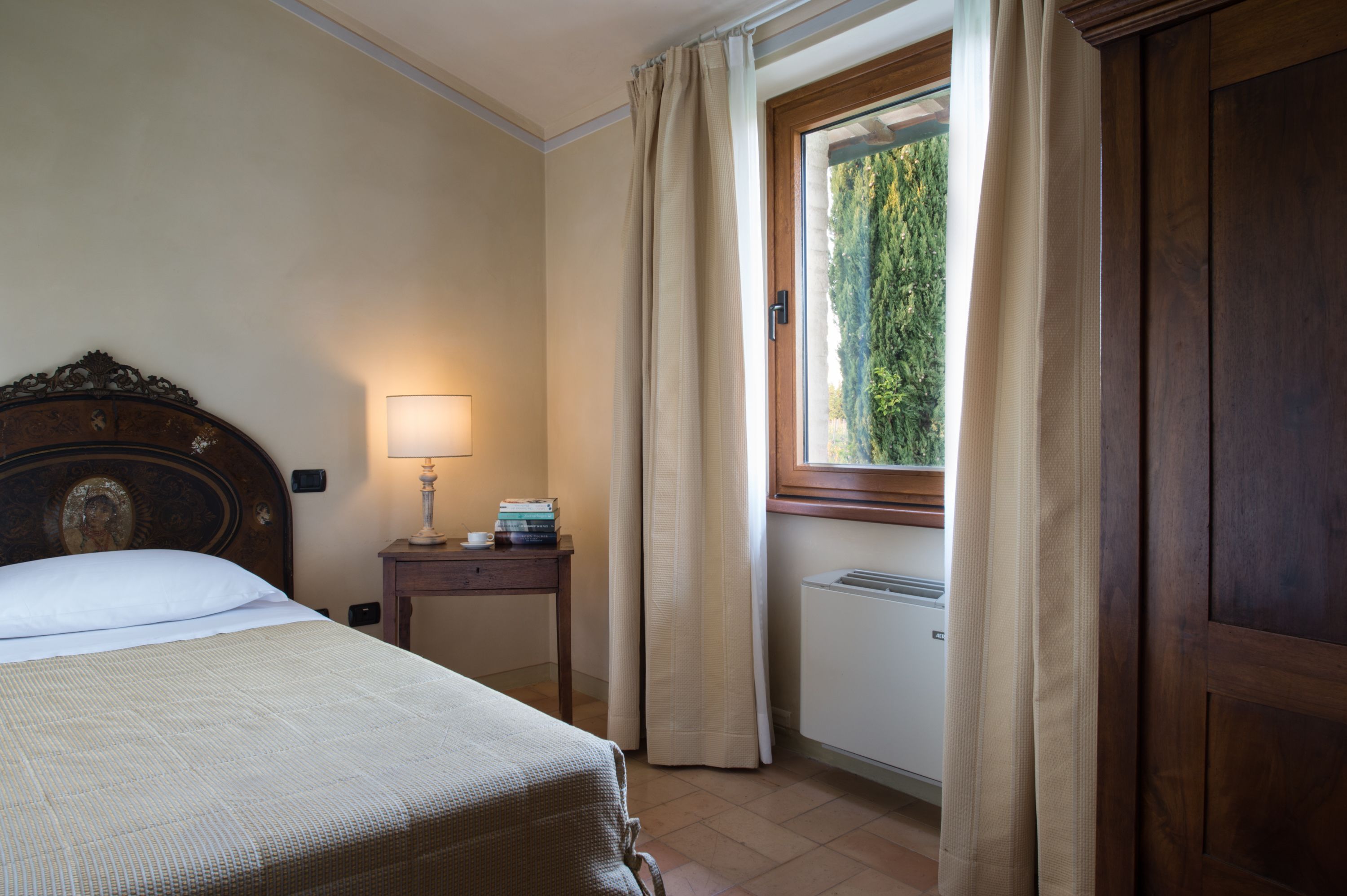A bedroom with a bed, a bedside table with a lamp, a window with curtains, and a radiator.