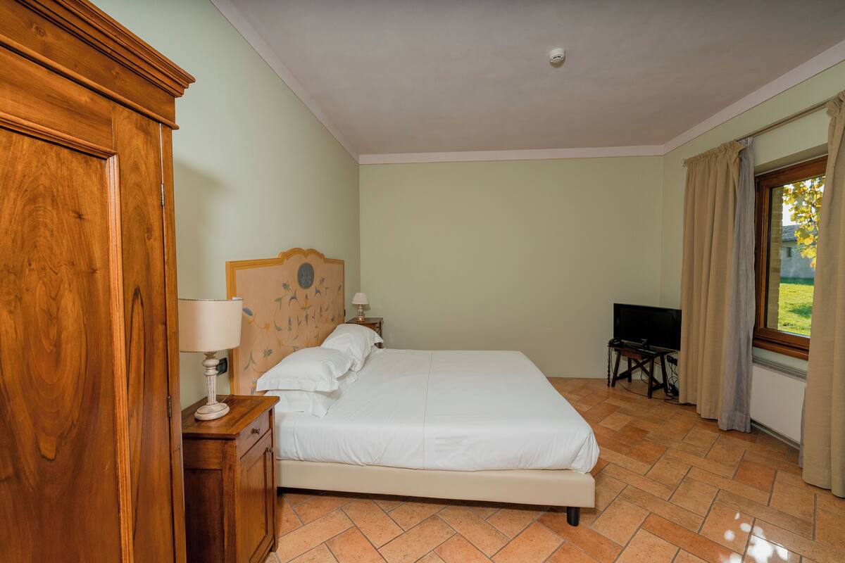 A bedroom with a wooden wardrobe, a bed with white linens, a nightstand with a lamp, and a television on a small table.