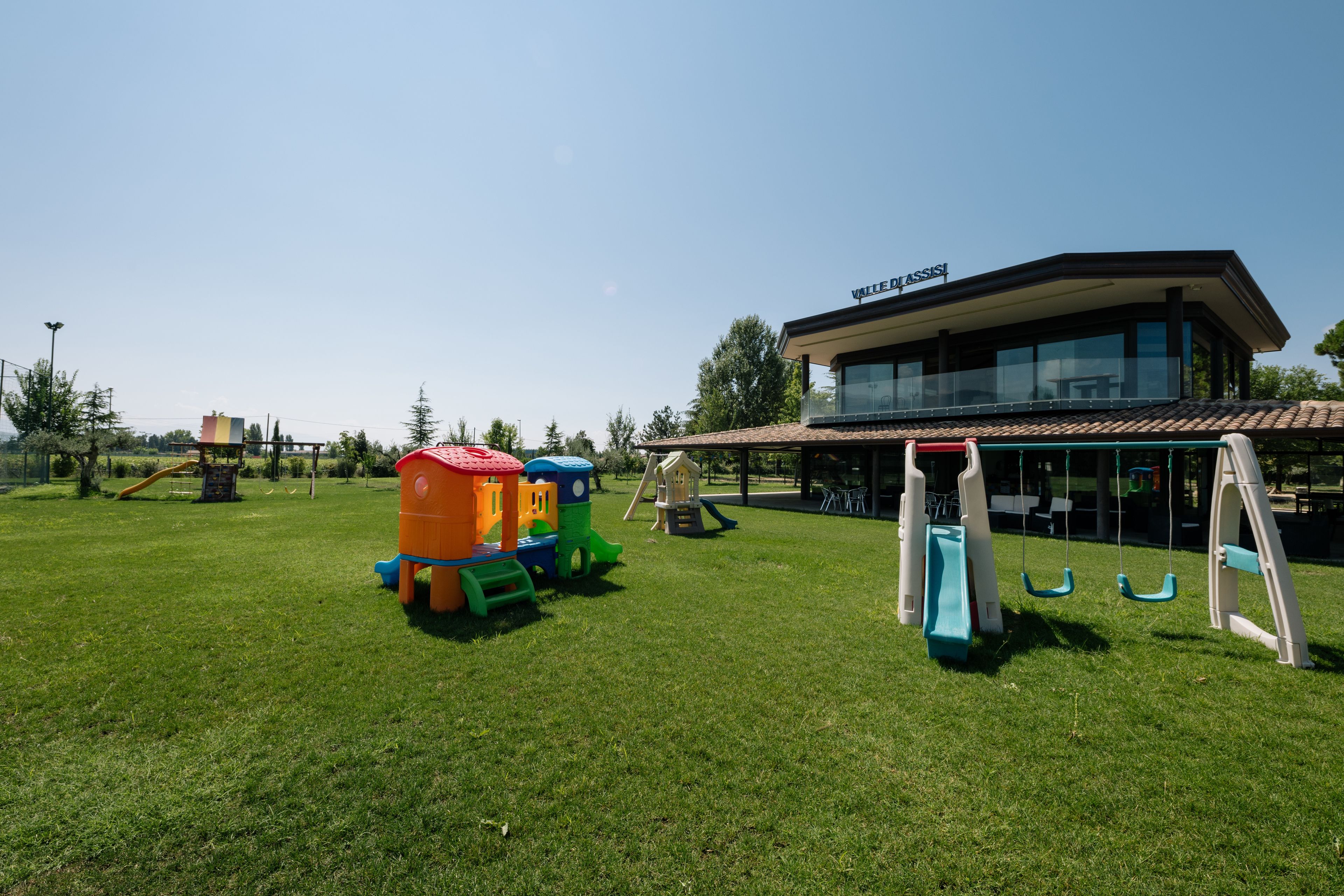 Un parque infantil con tobogán y columpios, un edificio con un letrero que dice "VILLAGE ASSI", y un cielo despejado.