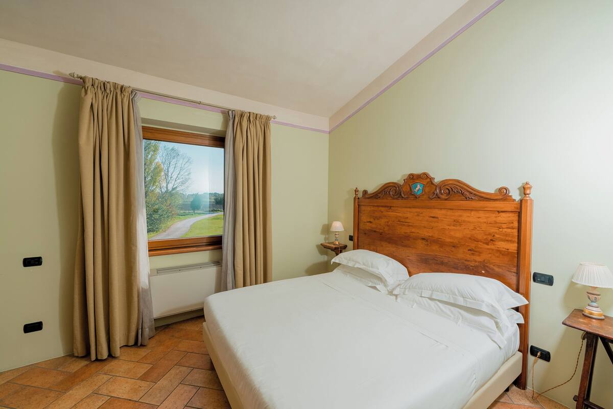 A bedroom with a wooden bed, beige curtains, a window with a view of a green landscape, and a small bedside table with a lamp.