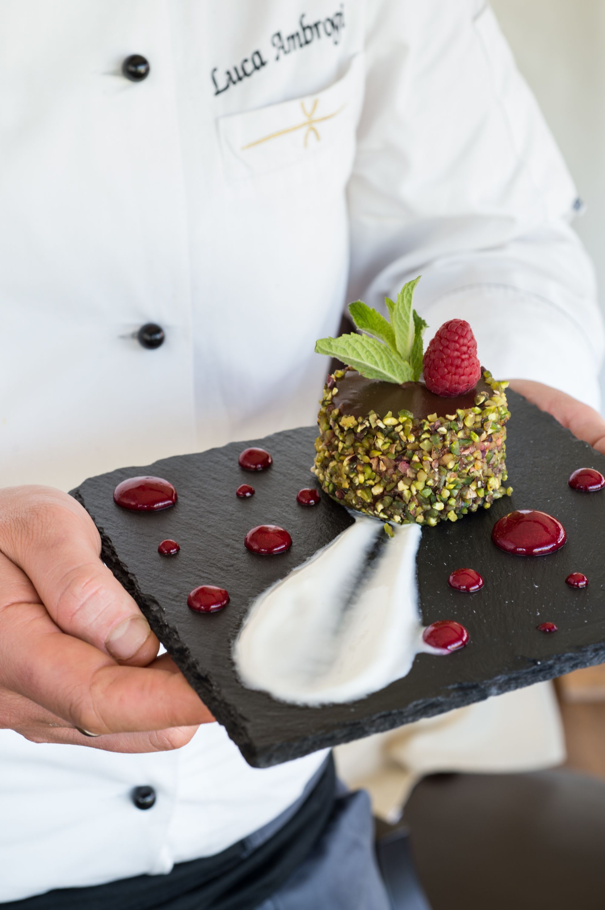 Un chef sosteniendo un postre con adorno de frambuesa y menta sobre un plato de pizarra negra.