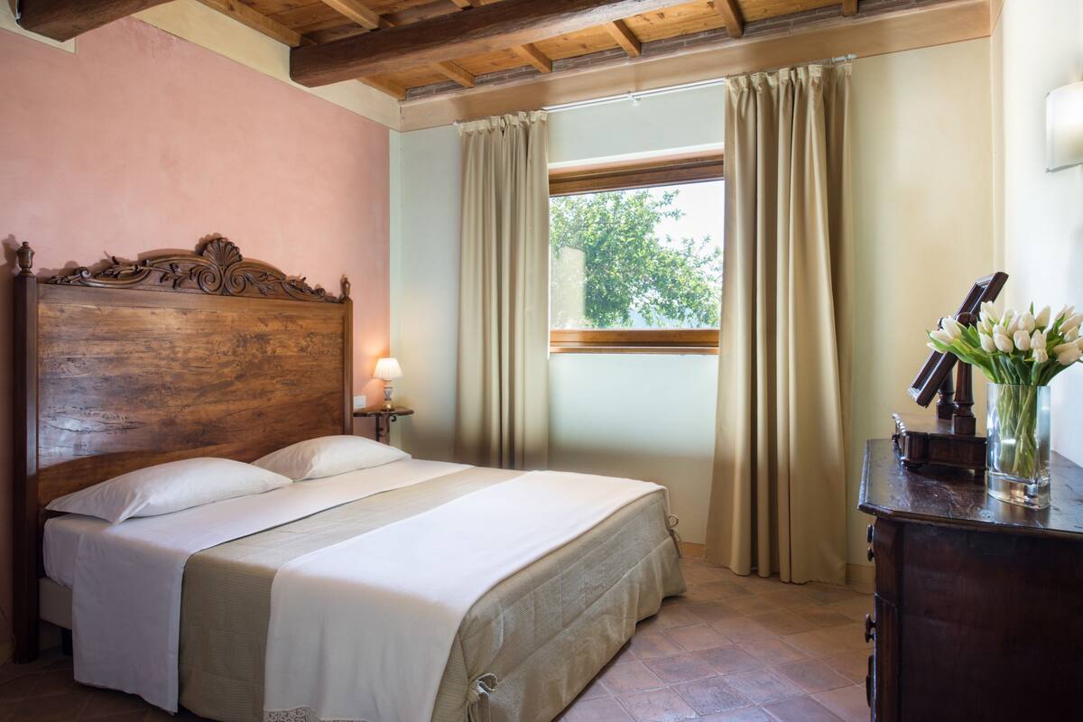 A bedroom with a wooden headboard, a bed with white linens, a nightstand with a lamp, a dresser, and a window with curtains.