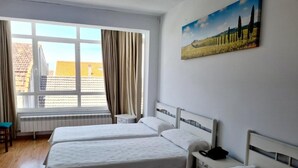 Habitación de hotel con tres camas, una ventana con cortinas, un radiador, una mesita y un cuadro en la pared.