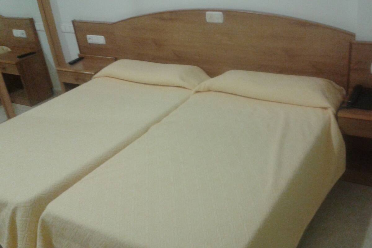 A twin bed with a wooden headboard and two pillows, a bedside table with a phone, and a small desk with a chair.