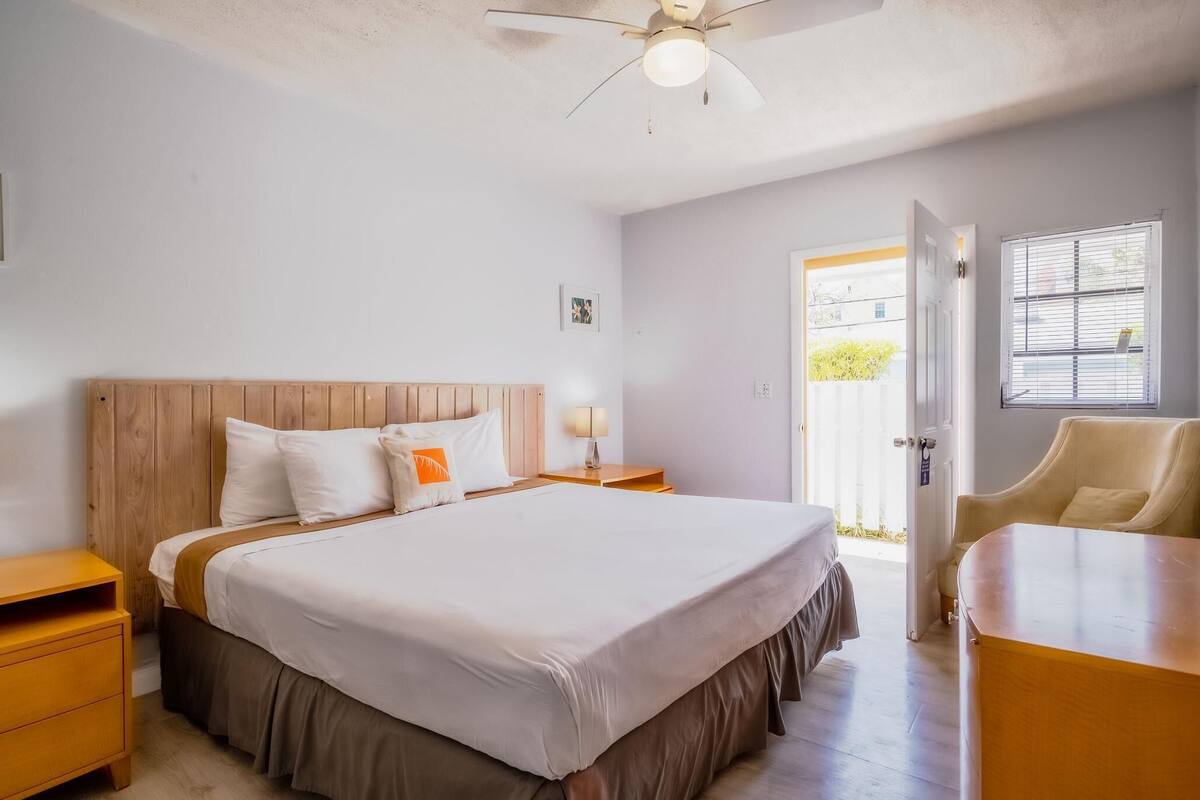 A bedroom with a wooden headboard, a bed with white bedding, a nightstand with a lamp, a wooden desk, and a chair.