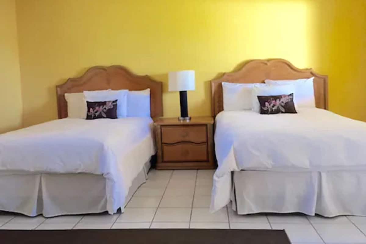 A hotel room with two beds, each with a wooden headboard, white bedding, and a nightstand with a lamp between them.