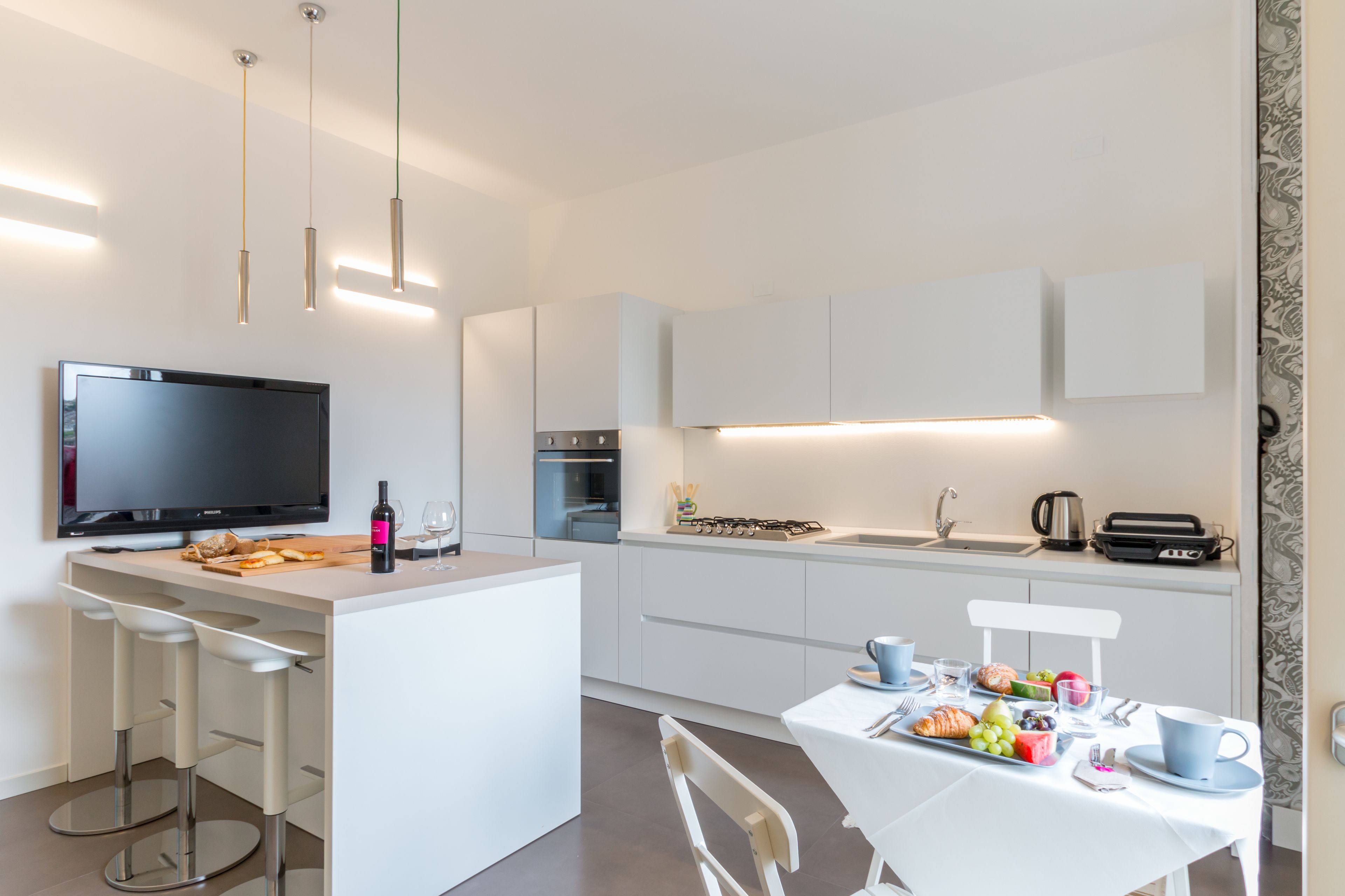 A modern kitchen with a central island, bar stools, a flat-screen TV, and a dining area with a table set for two.
