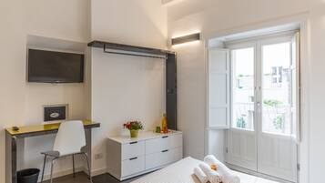 A bedroom with a bed, a desk, a chair, a wardrobe, and a window with a view of buildings.