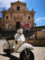Een witte Vespa scooter geparkeerd voor een historisch gebouw met versierde architectuur, een stopbord en een trap die naar de ingang leidt.