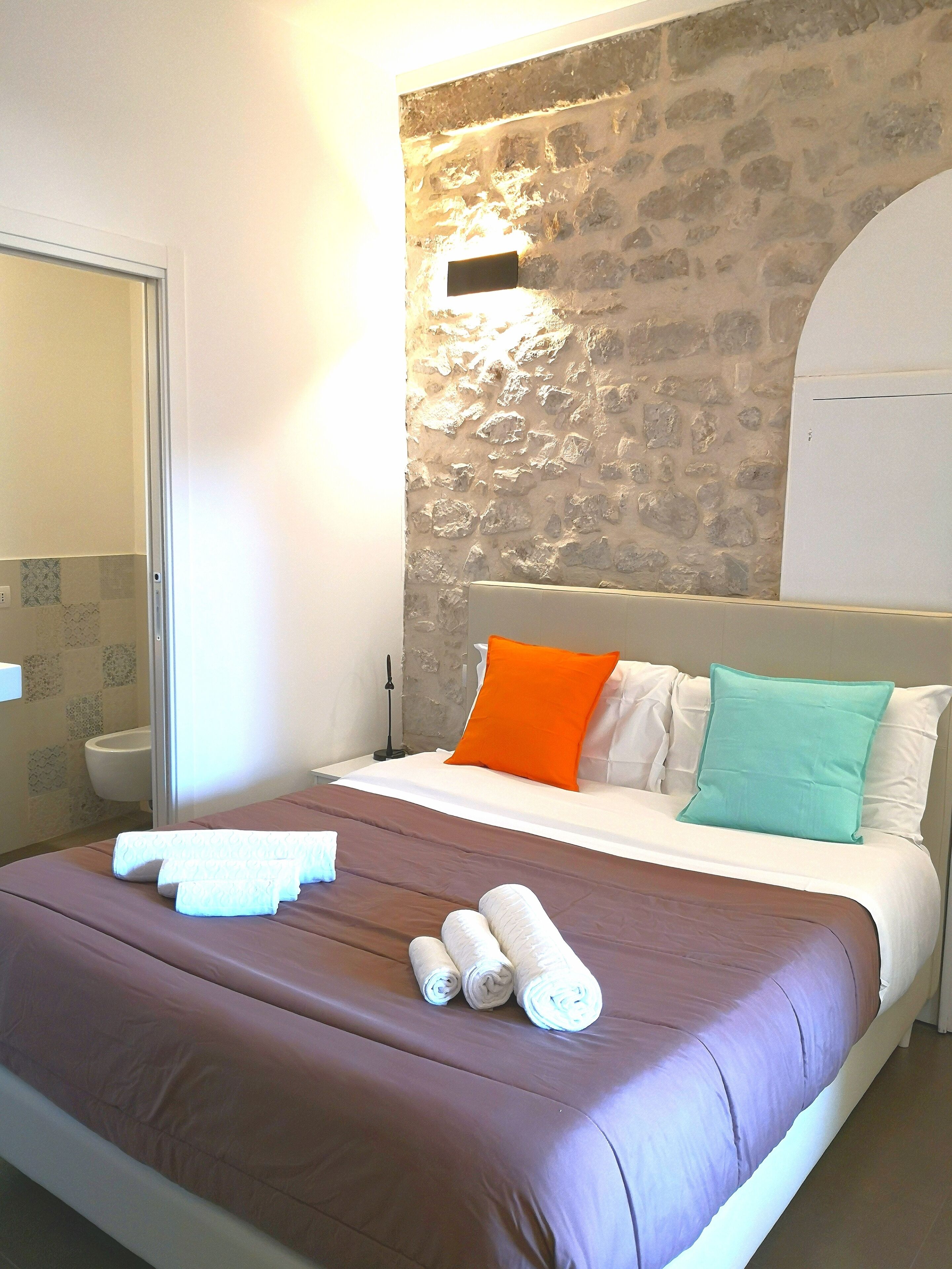 Habitación de hotel con una pared de acento de piedra, una cama con almohadas blancas y anaranjadas, y una cama bien hecha con toallas.