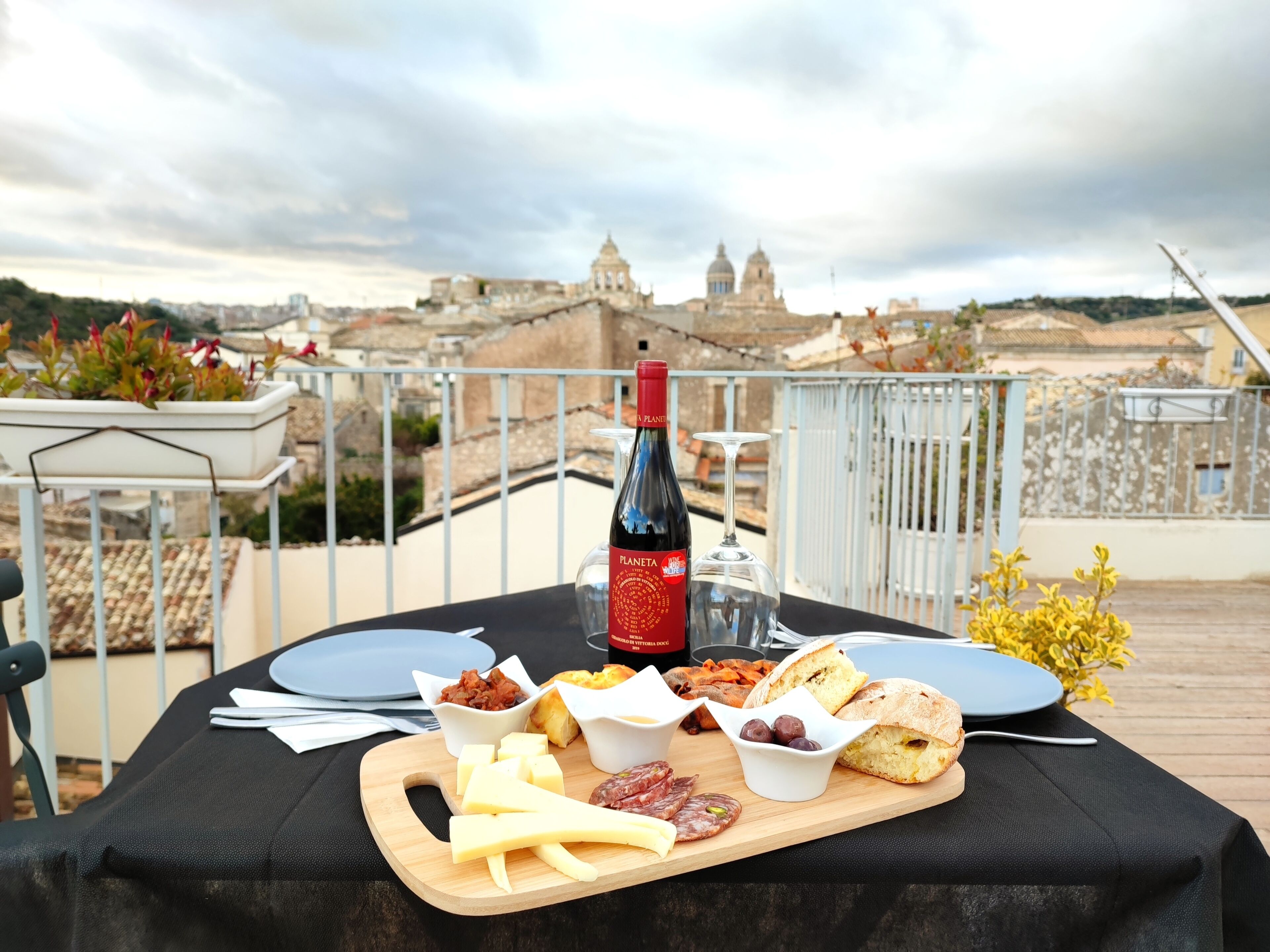 A table set for a meal with a bottle of wine, cheese, cured meats, olives, bread, and a view of a historic cityscape in the background.