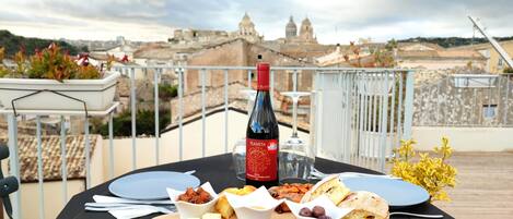 Una mesa preparada para una comida, con una botella de vino, queso, embutidos, aceitunas, pan y una vista de un paisaje urbano histórico al fondo.