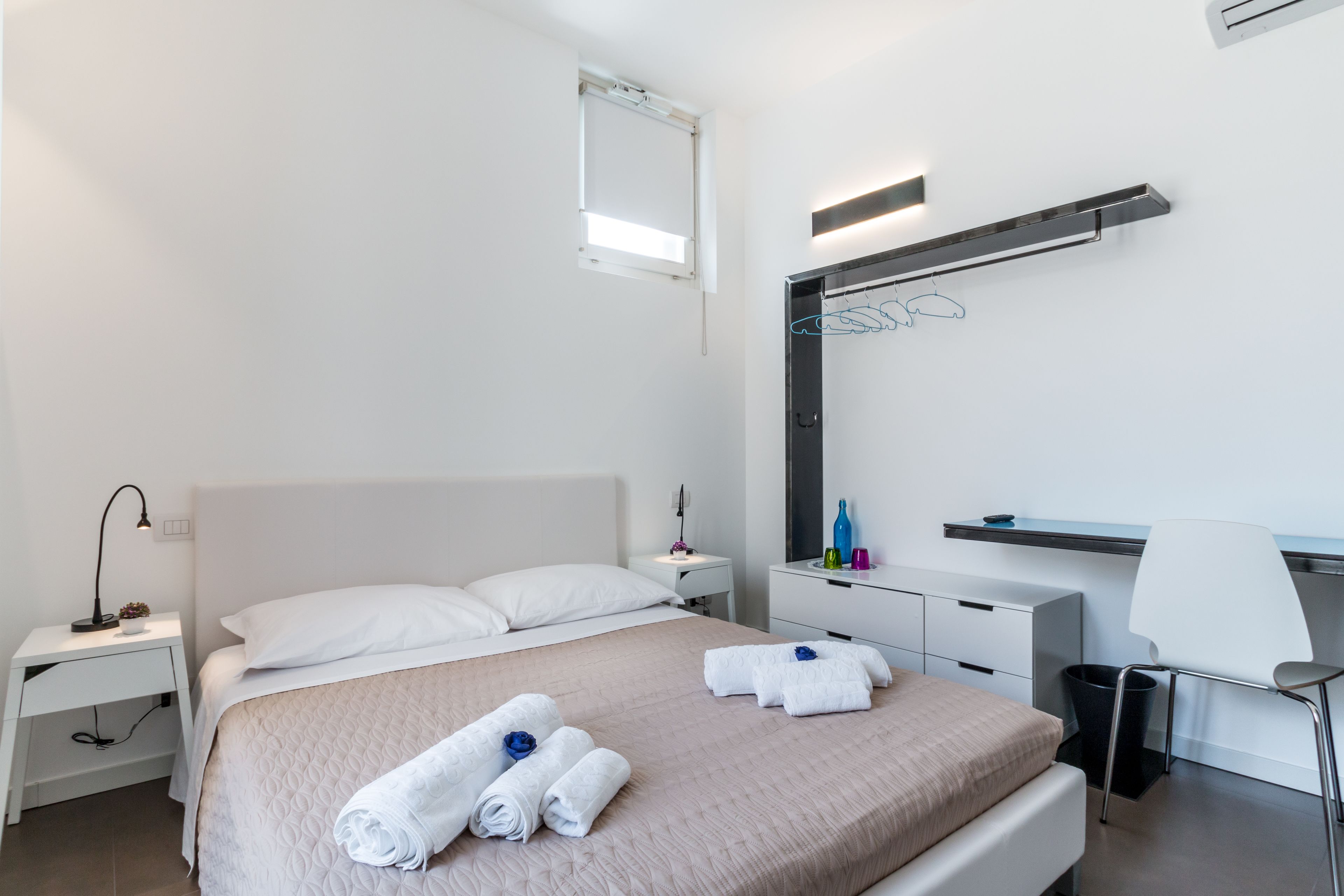 A modern hotel room with a neatly made bed, a bedside table with a lamp, a small desk with a chair, and a large mirror on the wall.