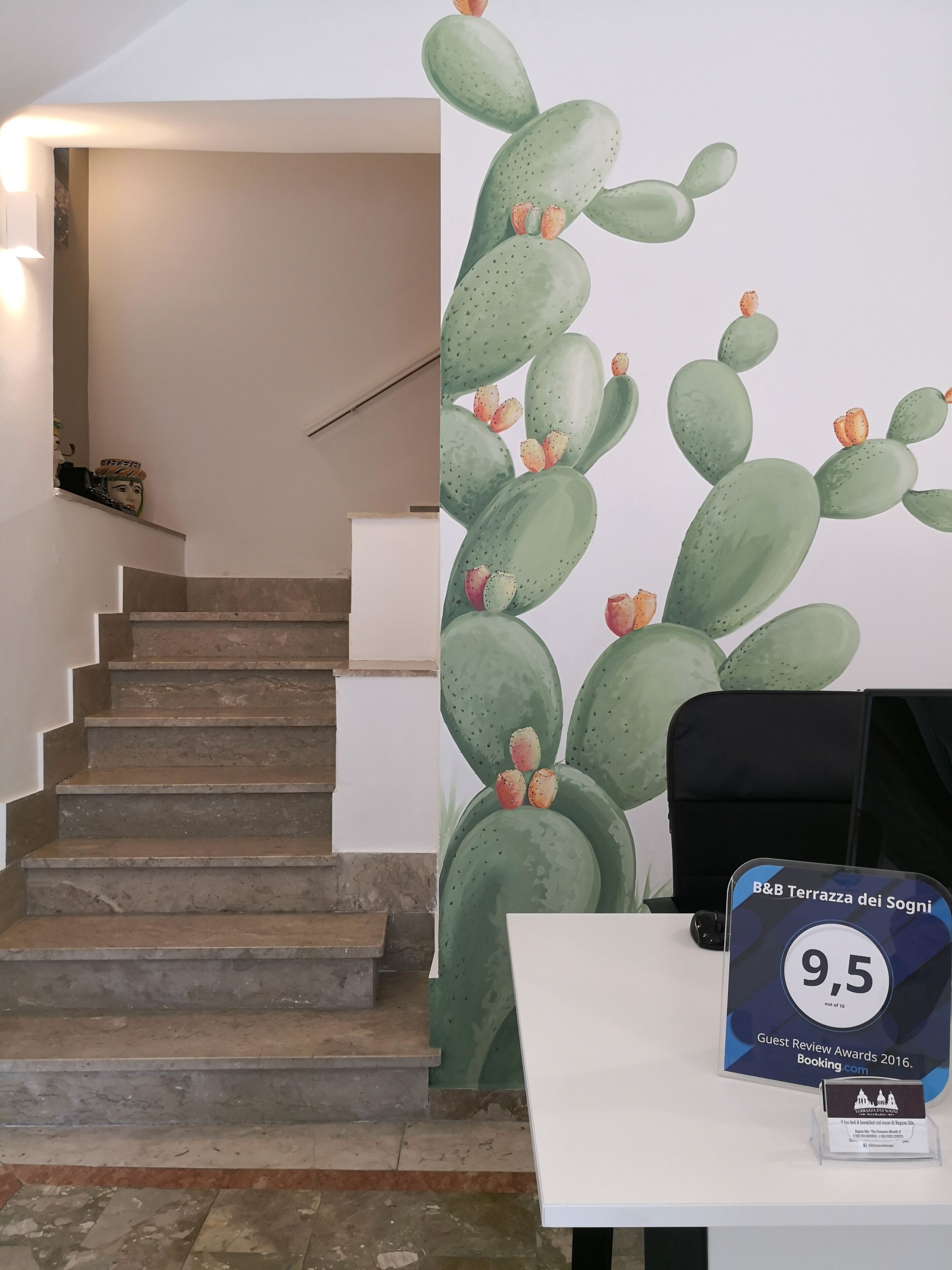A hotel interior with a staircase, a cactus wall mural, and a desk displaying a guest review award plaque.