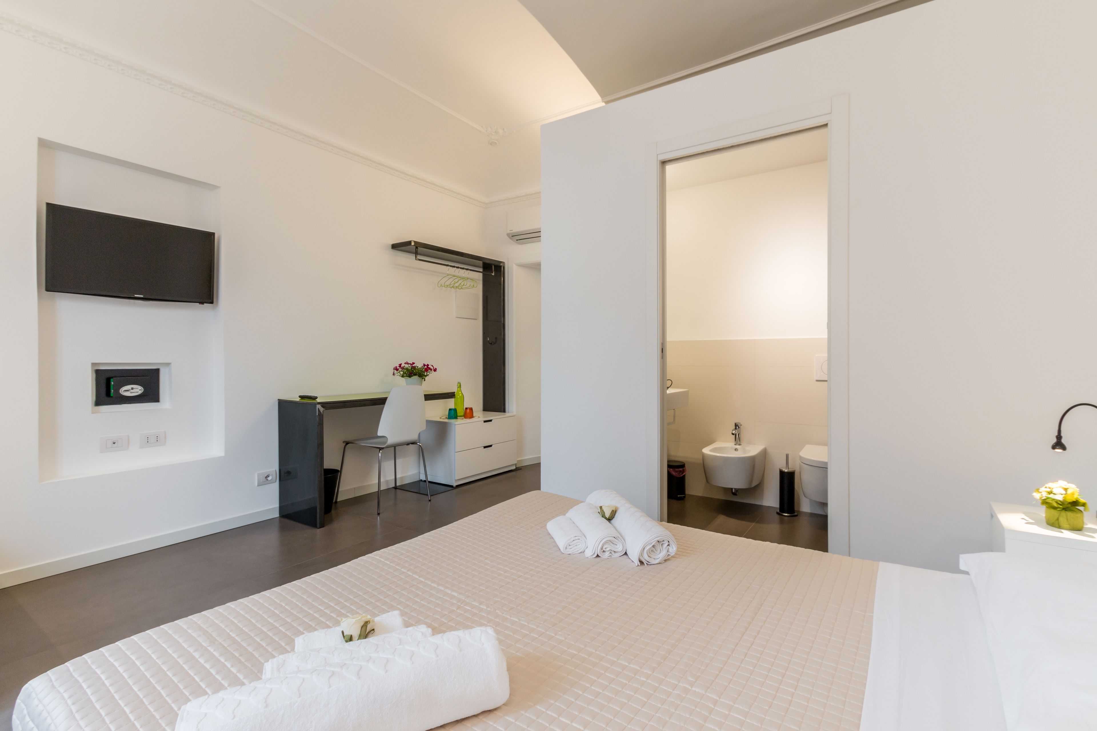 A hotel room with a large bed, a desk with a chair, a flat-screen TV mounted on the wall, and a bathroom with a sink and toilet.