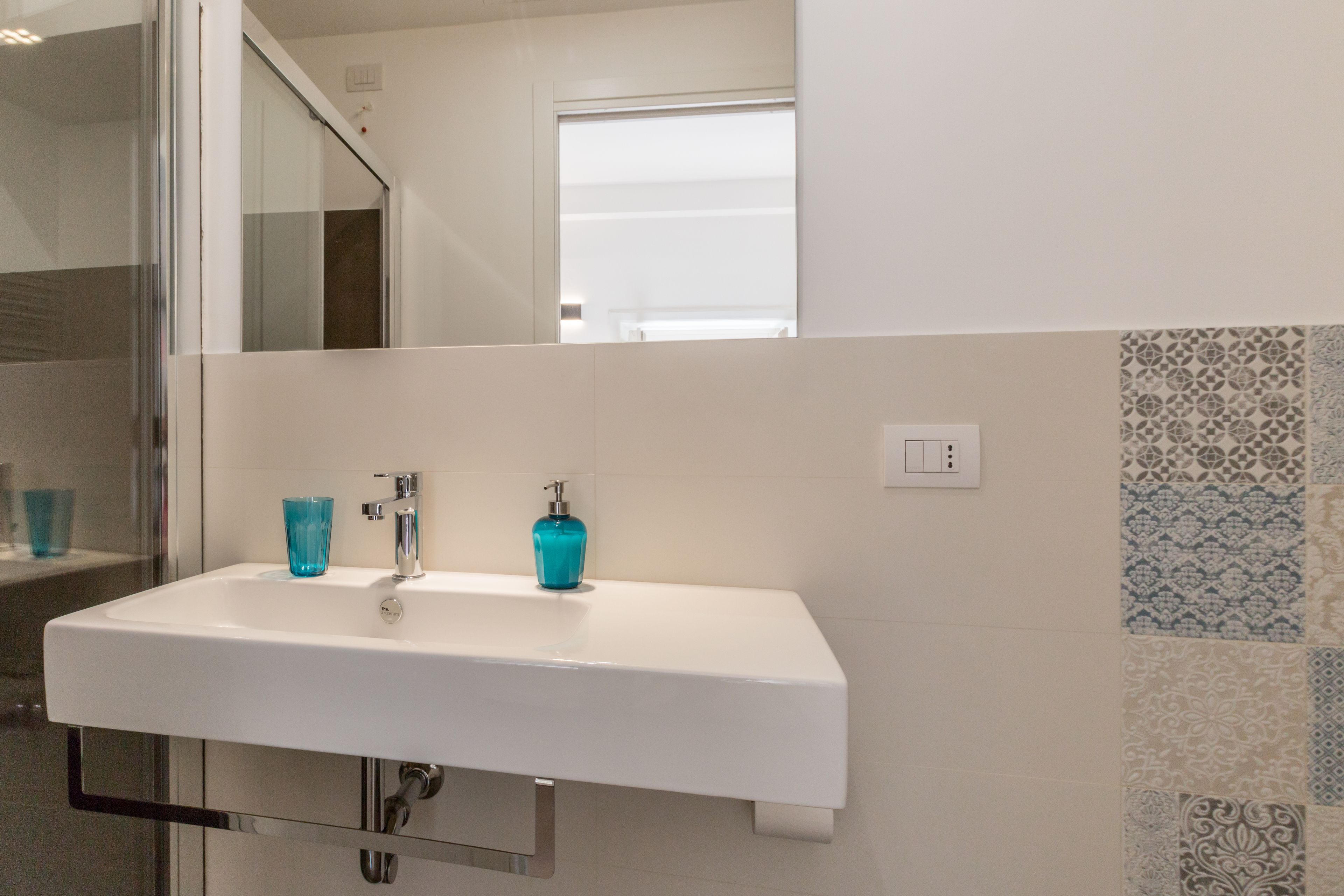 Un baño con un lavabo blanco, un vaso de vidrio azul y azulejos estampados en la pared.