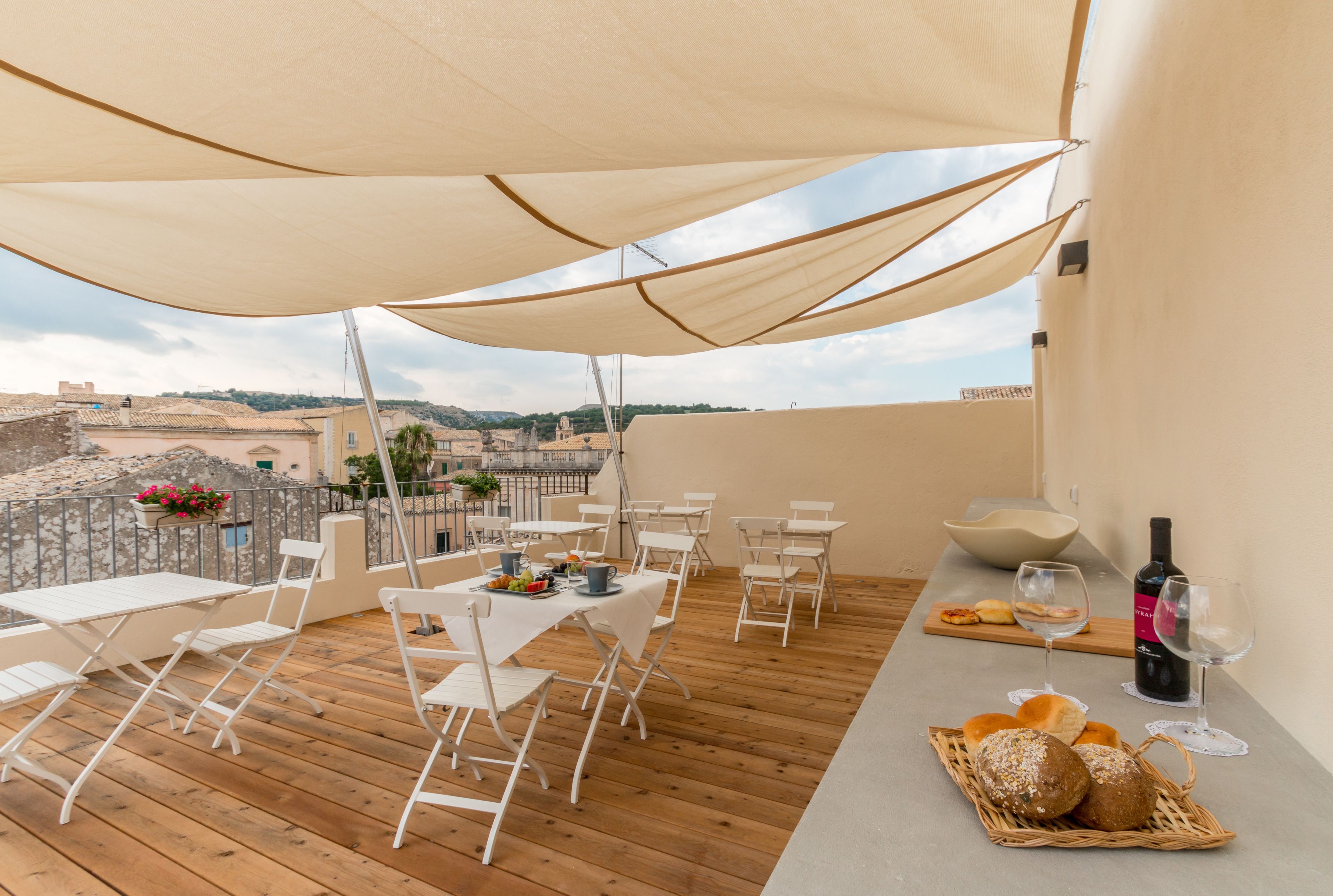 A terrace with white tables and chairs, a wicker basket with bread, a bottle of wine, and a bowl.