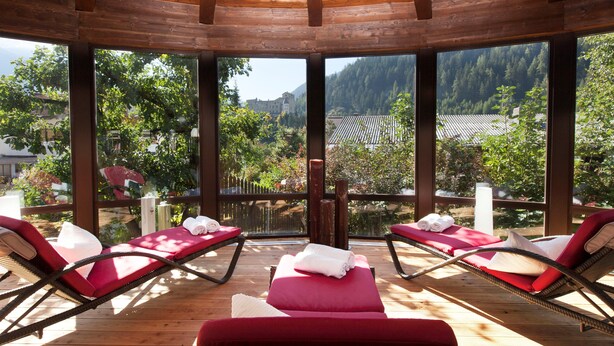 A sunlit outdoor lounge area with wooden flooring and red cushioned chairs, overlooking a forested valley and a castle. - Beauty & Wellness Hotel Tirolerhof (Nauders)