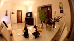 Uma sala de jantar com uma mesa posta para dois, uma televisão ao fundo e uma grande planta na parede.