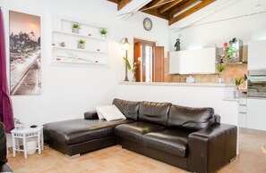 Un salon avec un canapé en cuir marron, une table d’appoint blanche, une petite table blanche, une étagère murale ornée de plantes, une horloge et un coin cuisine avec des placards et une cuisinière.