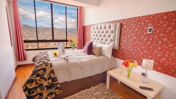 A bedroom with a large window, a bed with patterned bedding, a bedside table with a lamp and vase, and a view of a mountainous landscape.