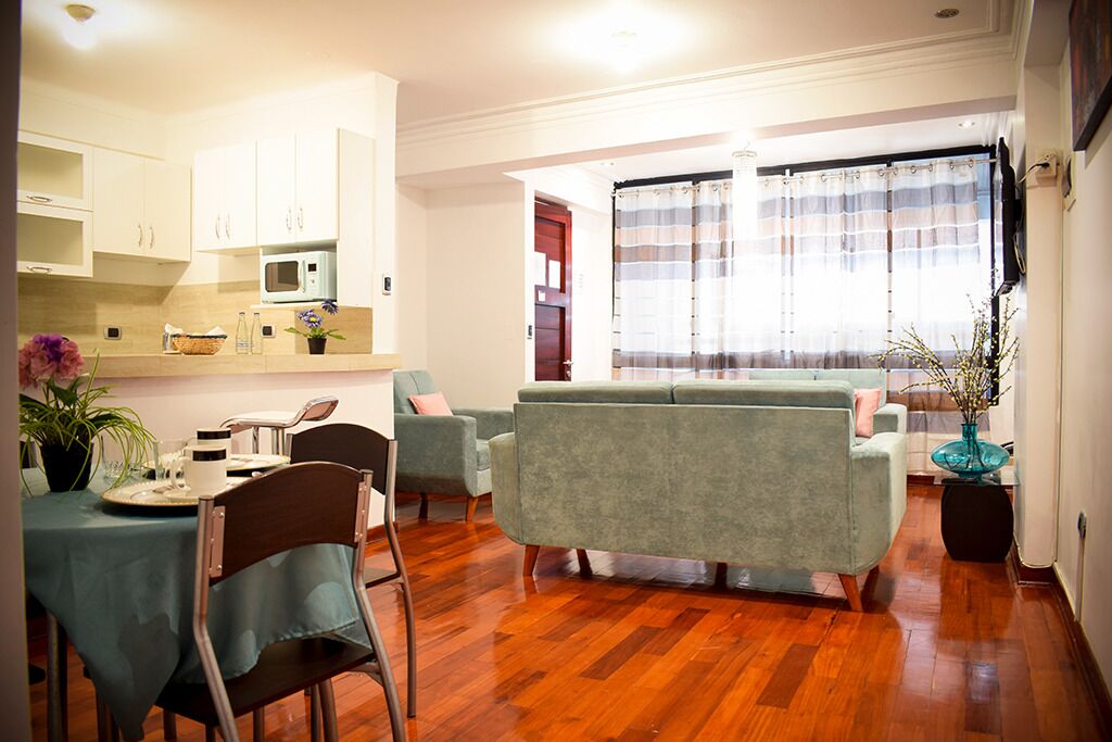 A modern living room with a sofa, a dining table with chairs, and a kitchen area with cabinets and a microwave.