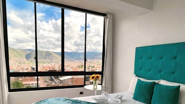 A hotel room with a large window offering a city view, a bed with a teal headboard, and a white quilt.