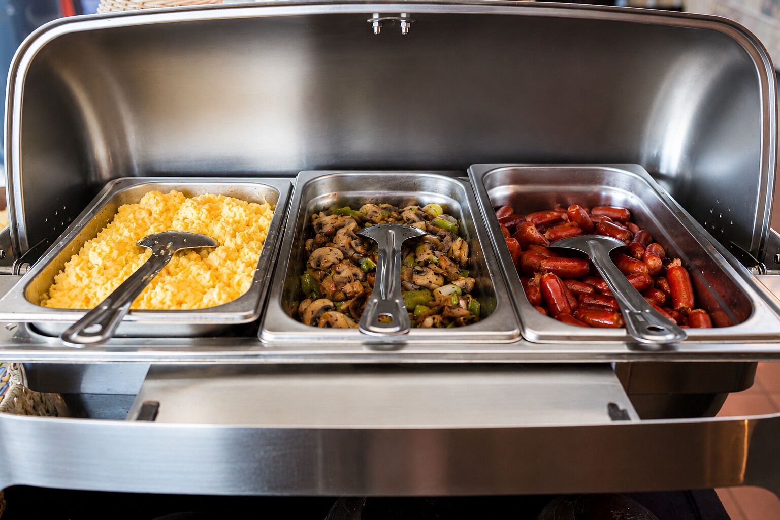 A buffet with three trays of food: scrambled eggs, sautéed mushrooms, and sausages.