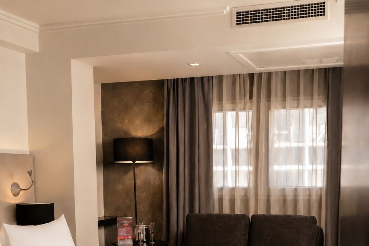 A hotel room with a bed, a sofa, a desk with a lamp, and a wall-mounted air vent.