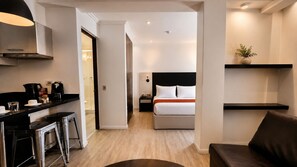 Ein modernes Hotelzimmer mit einer Küchenzeile, einem Bett mit weißer Bettwäsche, einem dunklen Kopfteil und einem kleinen Nachttisch.