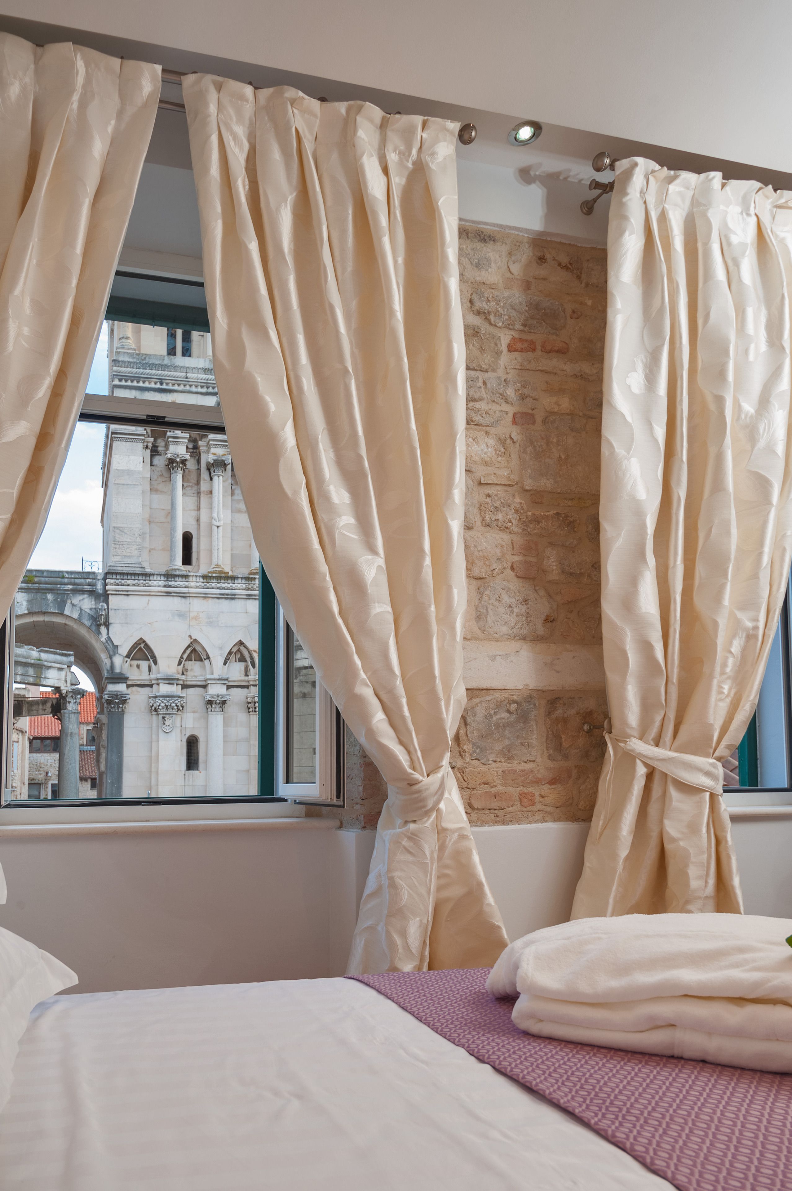 Quarto de hotel com uma cama, cortinas bege e vista para um edifício histórico pela janela.