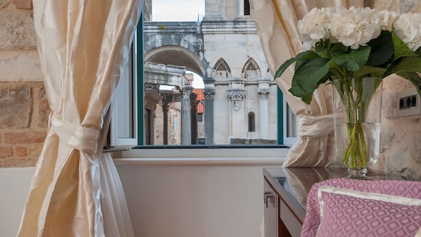 Una habitación con vistas a un edificio histórico, cortinas translúcidas, un florero con flores sobre una mesa y un cojín estampado.
