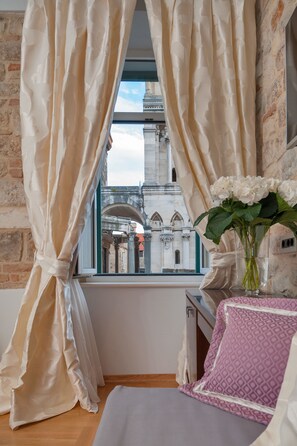 A room with a view of a historic building through sheer curtains, a vase with flowers on a table, and a patterned pillow. - Heritage Hotel Antique Split (Split)