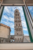 Une haute tour historique, dotée d’une horloge et d’un drapeau, que l’on aperçoit à travers une fenêtre à cadre vert.