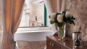 Una habitación con vistas a arquitectura histórica a través de una ventana, un florero con flores y un aparador de madera con una botella y vasos.