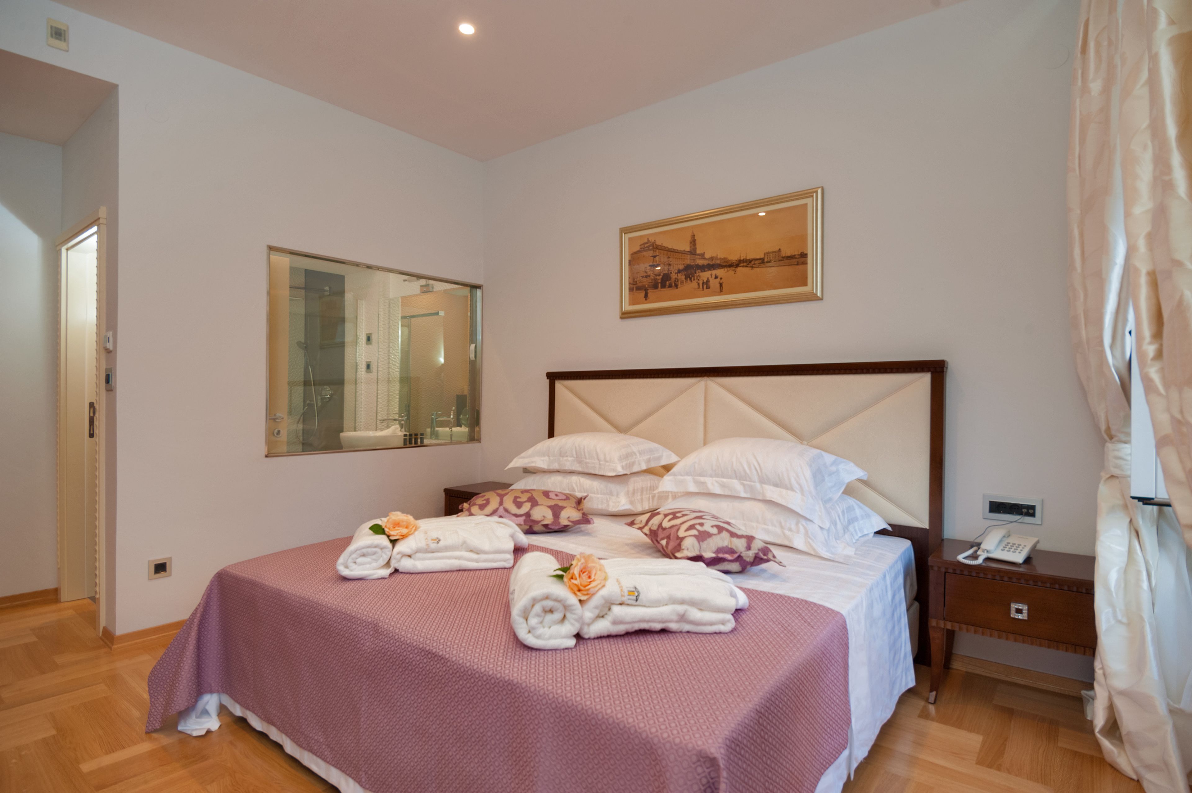 Una cama bien hecha con una colcha rosa, almohadas blancas y toallas con flores naranjas. En la pared, sobre la cama, hay un cuadro de un puente.