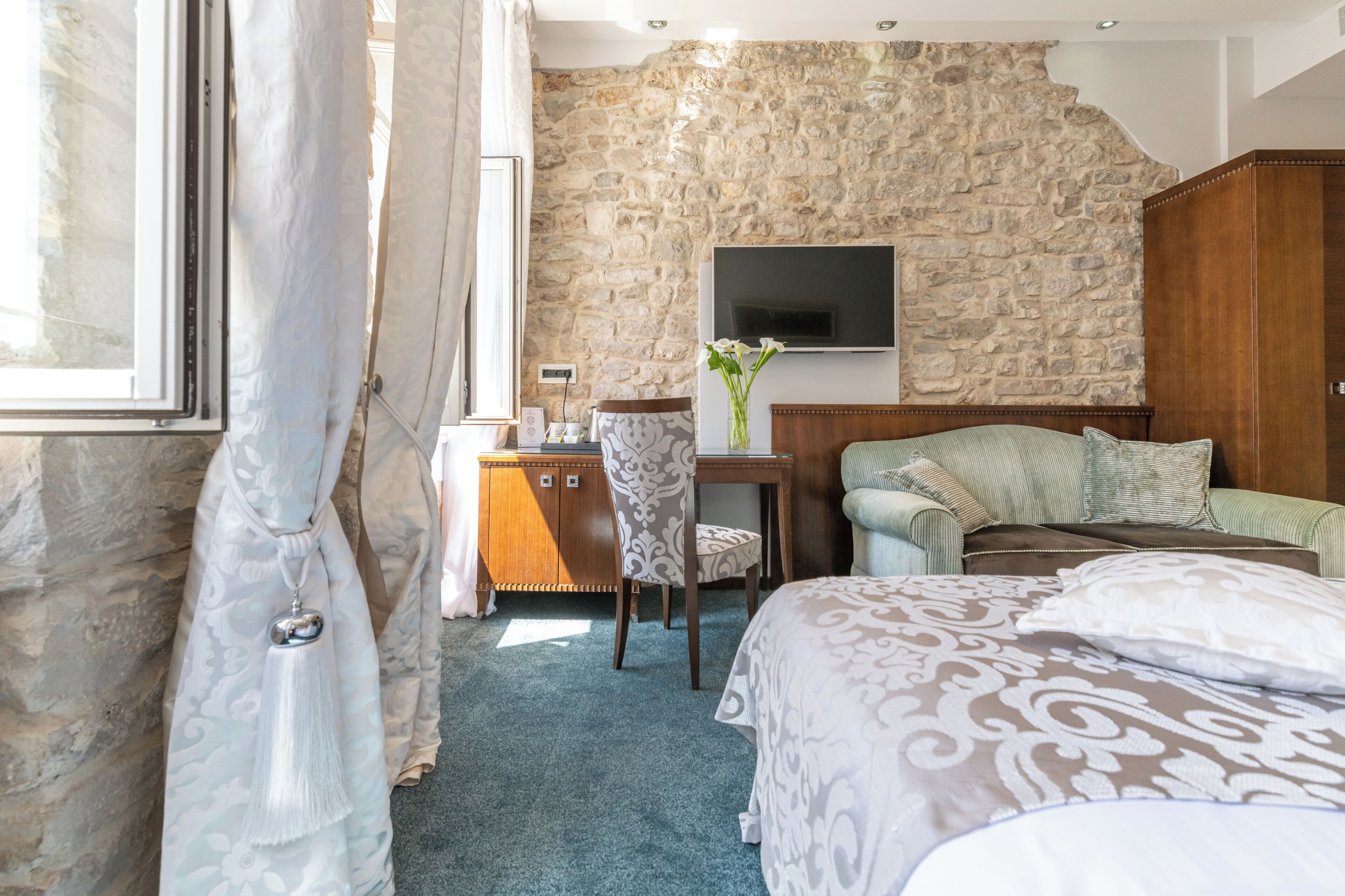 A bedroom with a stone wall, a bed with patterned bedding, a desk with a chair, a TV mounted on the wall, and a wardrobe.