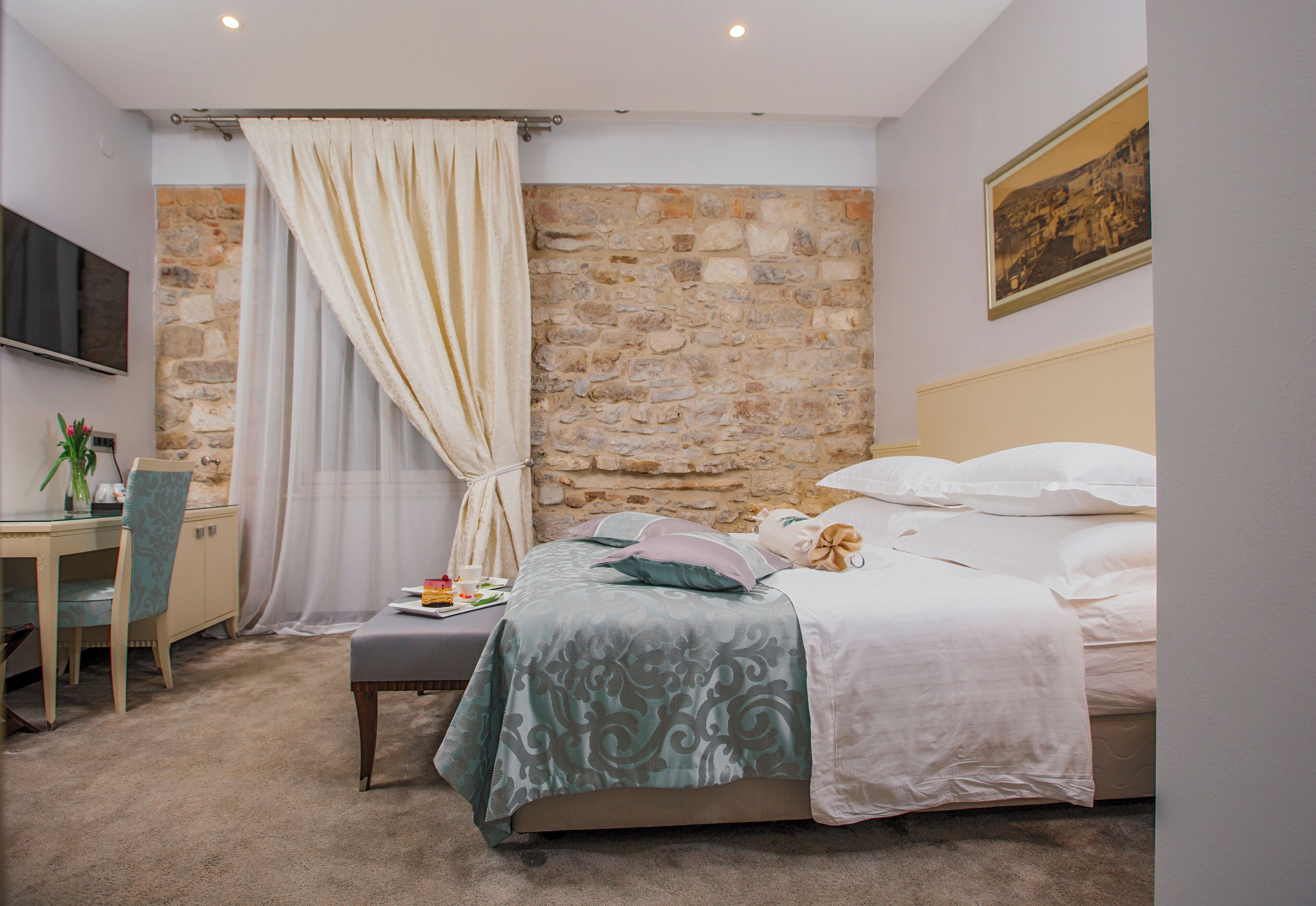 A bedroom with a stone wall, a bed with white bedding, a desk with a chair, and a television mounted on the wall.