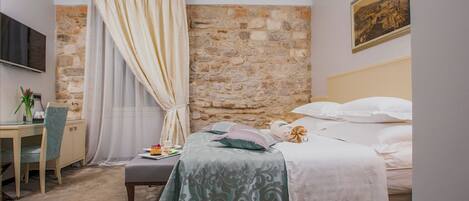 A bedroom with a stone wall, a bed with white bedding, a desk with a chair, and a television mounted on the wall.