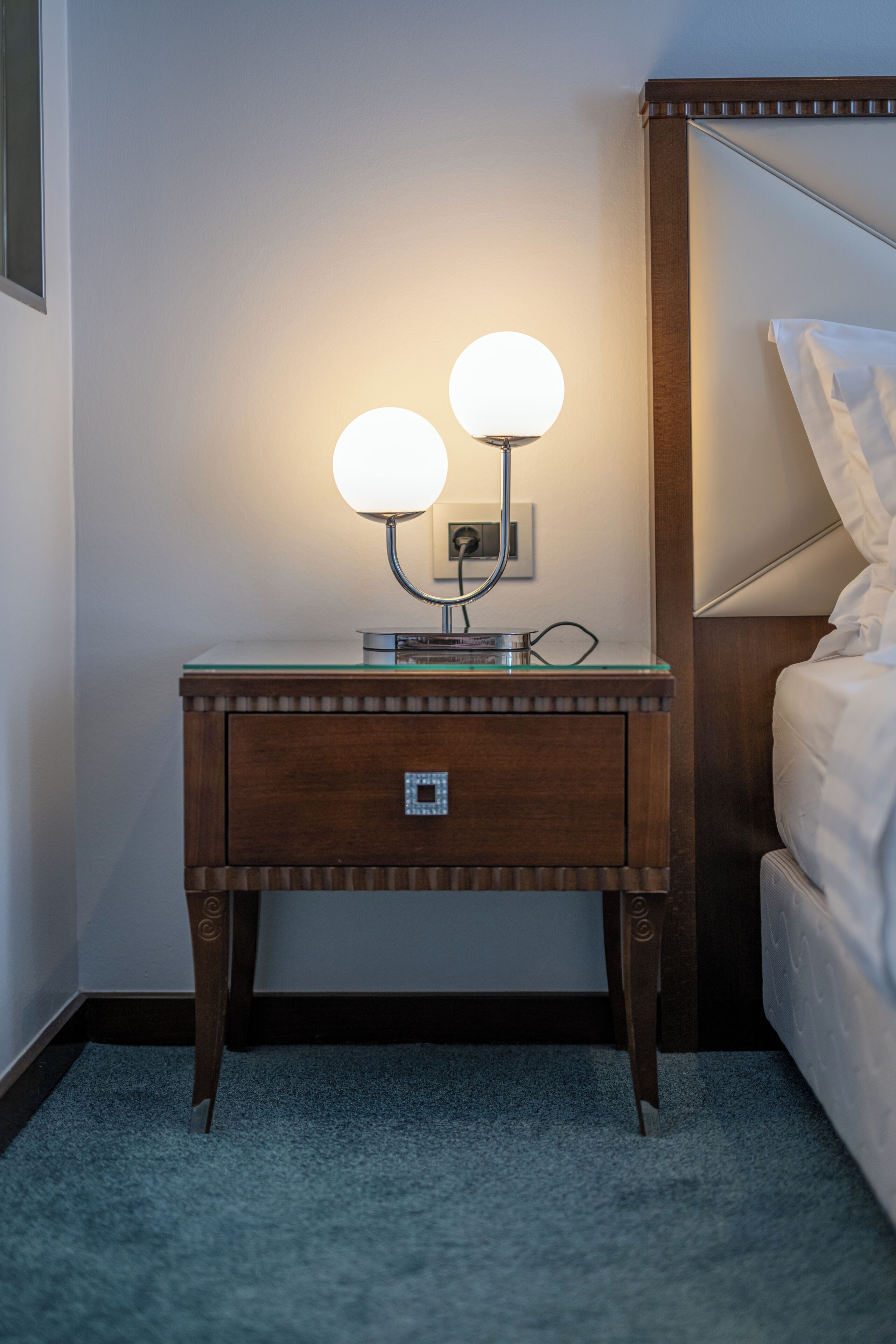 A wooden bedside table with two illuminated lamps, a glass top, and a drawer with a metallic handle.