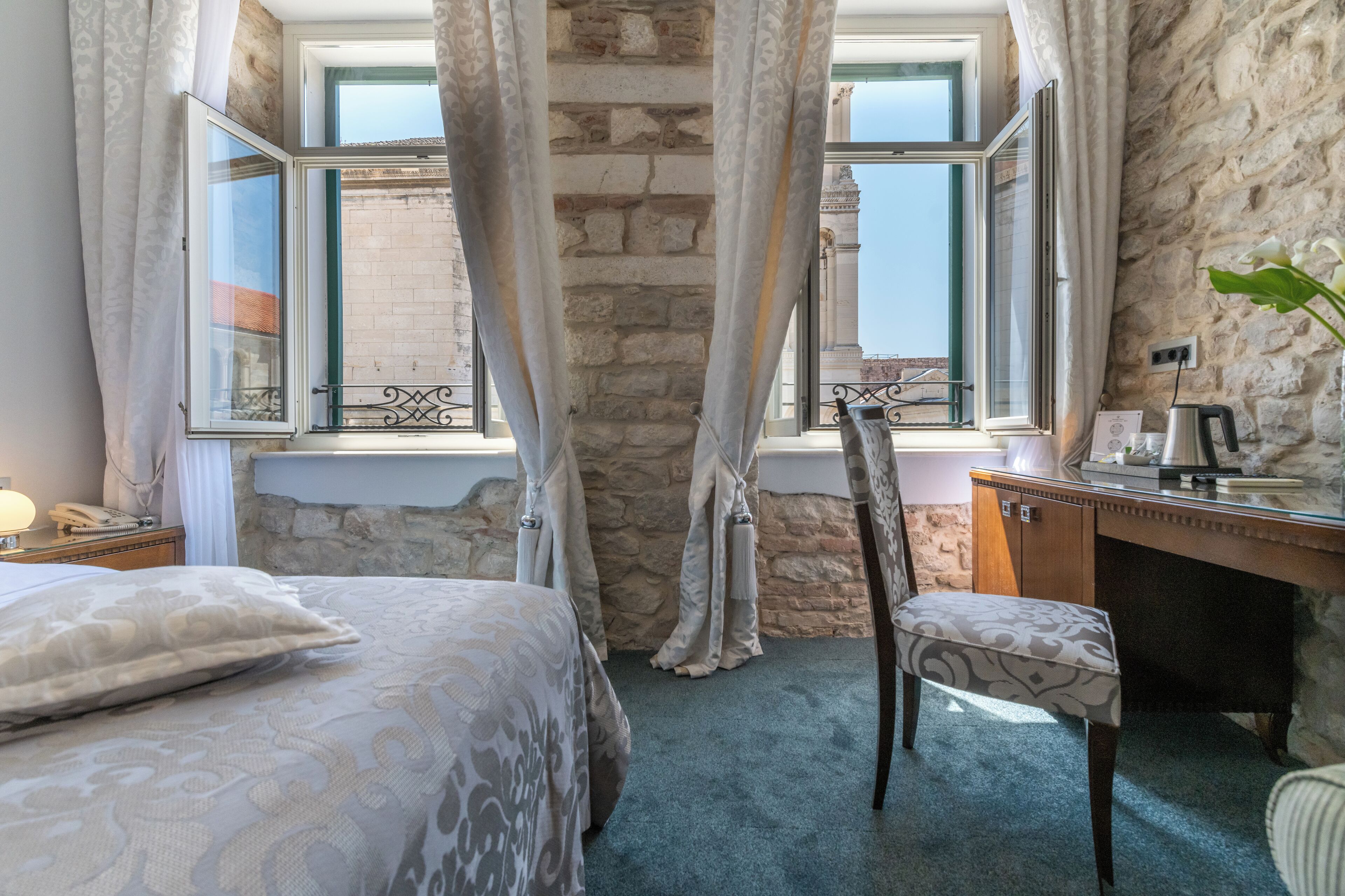 Un dormitorio con una cama, un escritorio y una silla. Hay cortinas en las ventanas y se aprecia una pared de piedra.