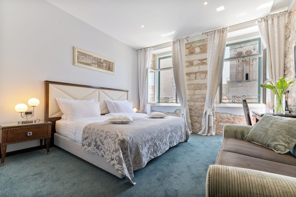 A hotel room with a large bed, a nightstand with a lamp, a sofa, and a view of a stone building through a window.