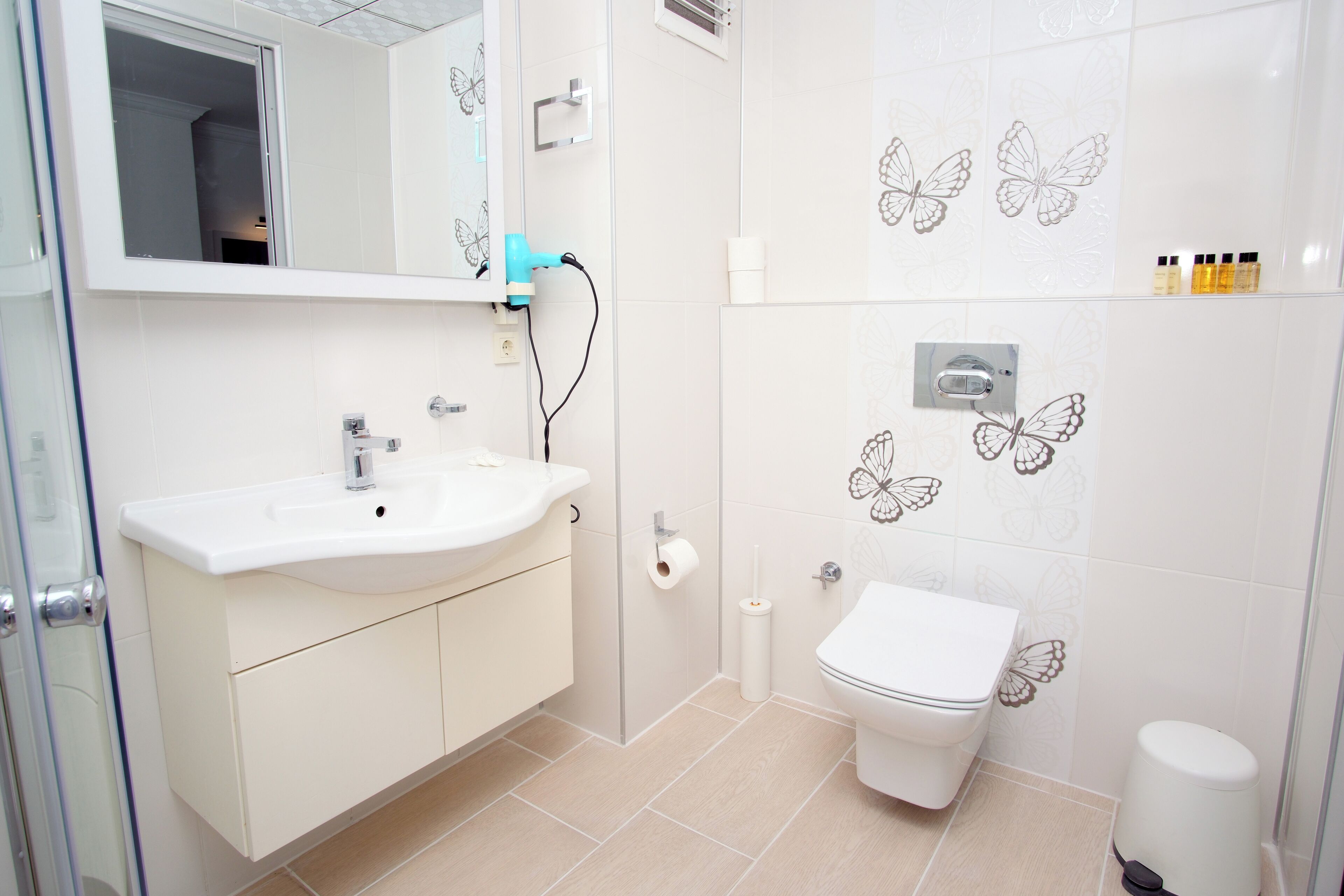 Un baño moderno con lavamanos, espejo y retrete blancos. Decoración con diseños de mariposas en las paredes y un estante pequeño con artículos de aseo.