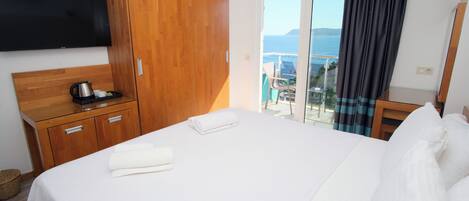 A hotel room with a bed, a wooden wardrobe, a flat-screen TV, a coffee maker, and a view of the sea through a sliding glass door.